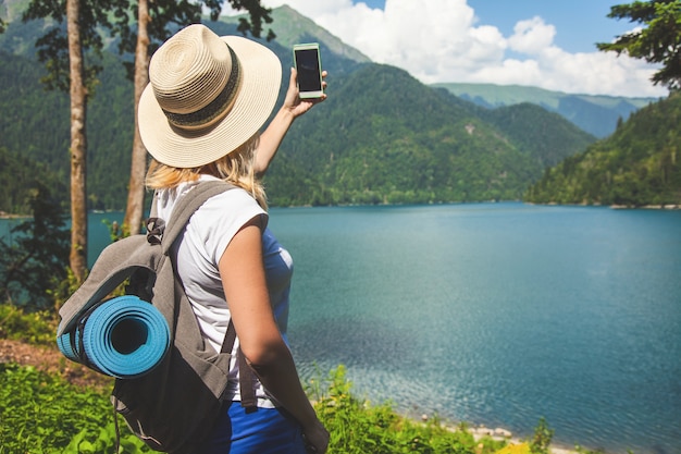 Beautiful girl traveler in a hat stands on a lake and takes pictures on a background of mountains