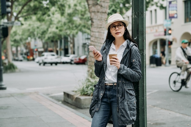 Beautiful girl walking around the city with a cup of coffee smiling. office lady relying on street lamp using mobile phone waiting for friends hold hot tea after work. man riding bike on background.