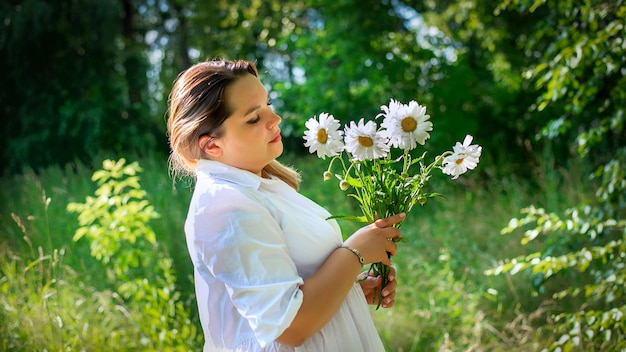 A beautiful girl walks in the forest with daisies at sunset of the day.