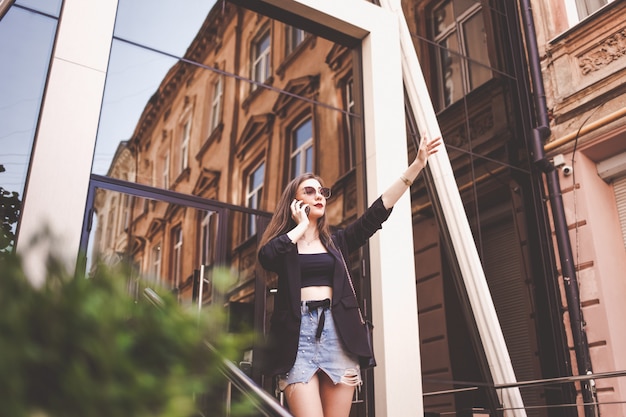 Beautiful girl waving. Stylish young woman talking on the phone and stops a taxi