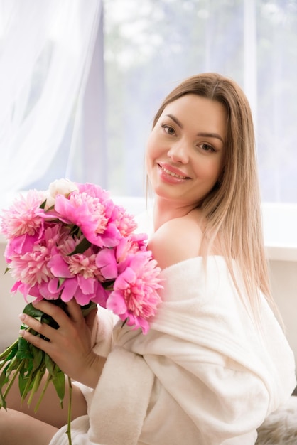 Beautiful girl in a white bathrobe with a bouquet of pink peonies near the bath