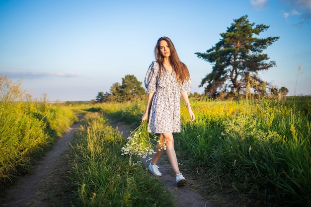 Beautiful girl in a white dress with a bouquet is walking along the road in the fields