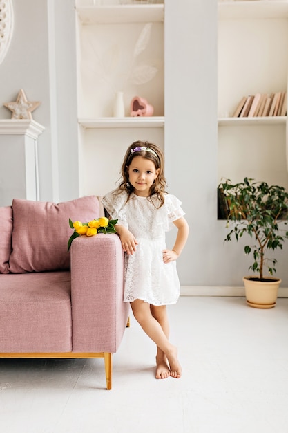 A beautiful girl in a white dress with a bouquet of tulips is standing next to her on the sofa in the room.