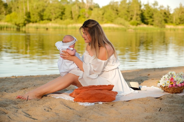 A beautiful girl in a white dress with a newborn baby is sitting on the beach on the sand