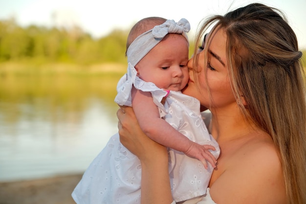 A beautiful girl in a white dress with a newborn baby is sitting on the beach on the sand