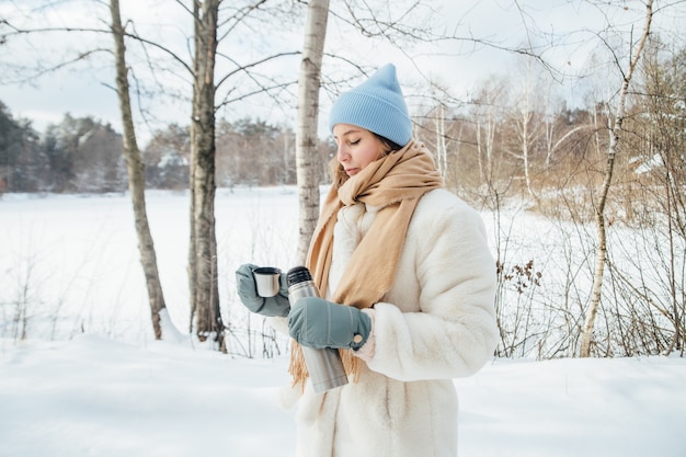 Beautiful girl in a white fur coat pours warm tea in winter in a snowy forest