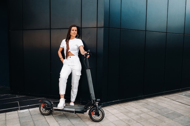 Beautiful girl in white stylish clothes on an electric scooter in the city.