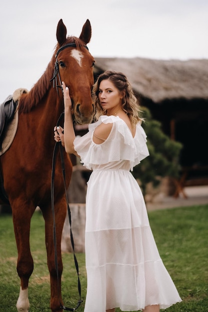 Beautiful girl in a white sundress next to a horse on an old ranch