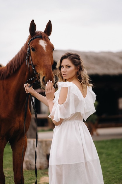 Beautiful girl in a white sundress next to a horse on an old ranch