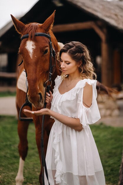 Beautiful girl in a white sundress next to a horse on an old ranch