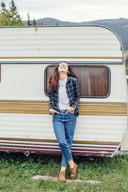 Beautiful girl with backpack traveling Girl standing near trailer traveling in mountains Woman in glasses traveling by car Traveler Girl thumbs up