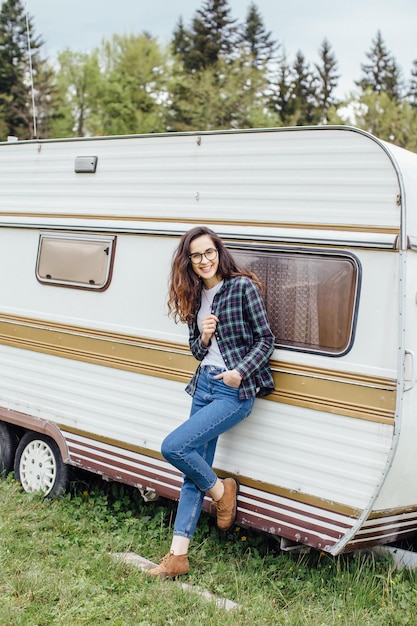 Beautiful girl with backpack traveling Girl standing near trailer traveling in mountains Woman in glasses traveling by car Traveler Girl thumbs up