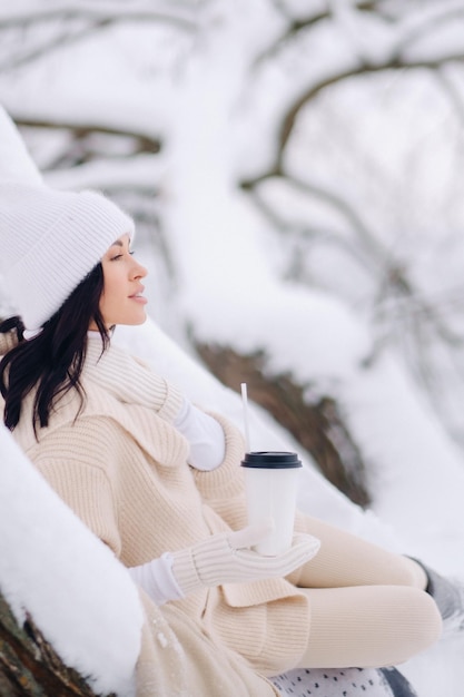 A beautiful girl with a beige cardigan and a white hat enjoying drinking tea in a snowy winter forest near a lake