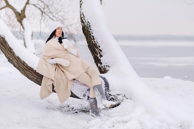 A beautiful girl with a beige cardigan and a white hat enjoying drinking tea in a snowy winter forest near a lake