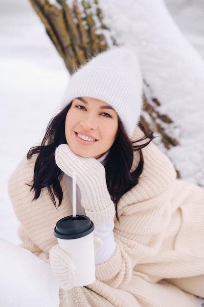 A beautiful girl with a beige cardigan and a white hat enjoying drinking tea in a snowy winter forest near a lake