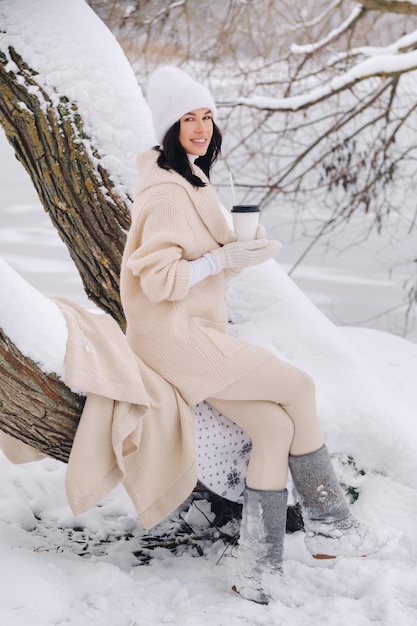 A beautiful girl with a beige cardigan and a white hat enjoying drinking tea in a snowy winter forest near a lake