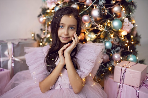 Beautiful girl with big eyes in a pink dress is sitting on the floor by the Christmas tree