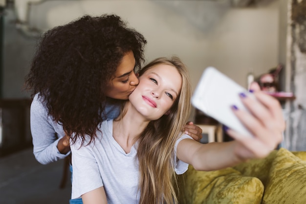 Beautiful girl with blond hair dreamily taking photos on cellphone while pretty african american girl kissing her in cheek while spending time together at home