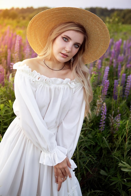 a beautiful girl with blonde hair in elegant white clothes and a straw hat on a summer field