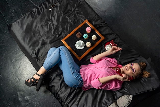 Beautiful girl with bright makeup in pink shirt and jeans lies on black bed Cupcakes with colored cream and coffee cup