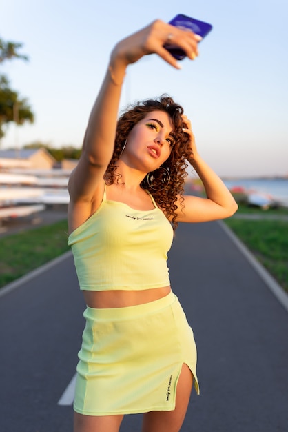 Beautiful girl with curls in a green skirt and top takes a selfie in the rays of the evening sun