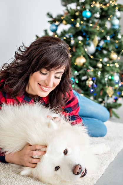 Beautiful girl with curls in a red-checked shirt laughs hugs a Samoyed husky that licks on the cheek