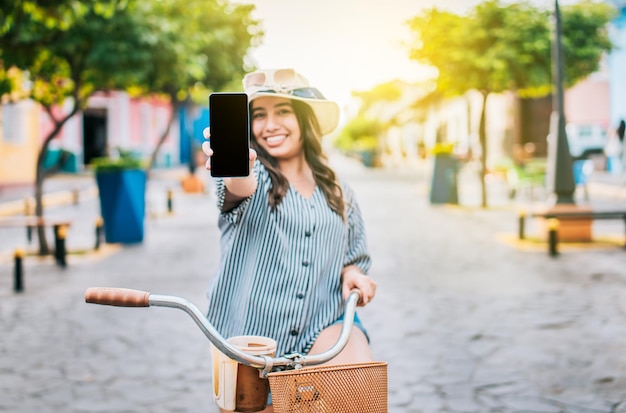 Beautiful girl with hat on bicycle showing cell phone screen on the street Smiling tourist on bicycle showing cell phone screen on the street