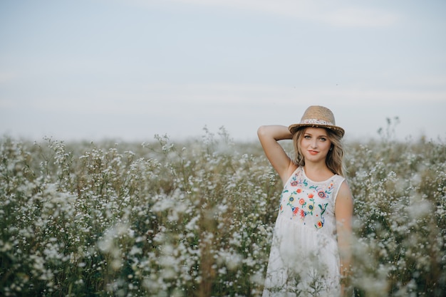 Beautiful girl with a hat in her hand walks in a field with field flowers and smiles sincerely