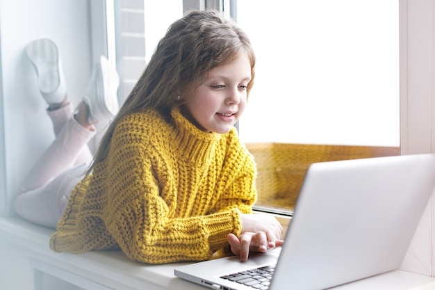 A beautiful girl with a laptop at home at the window looks at the screen and smiles. The girl at home communicates on the Internet. Kids distance learning.