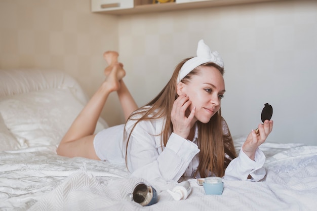 Beautiful girl with long hair in bed takes care of her skin. Using cosmetic creams