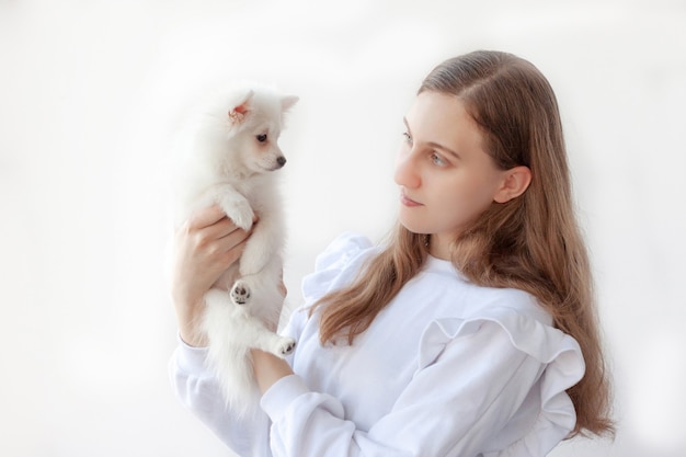 A beautiful girl with long hair in a white blouse holds a small, white Pomeranian dog in her hands and looks at it. The concept of animal welfare.