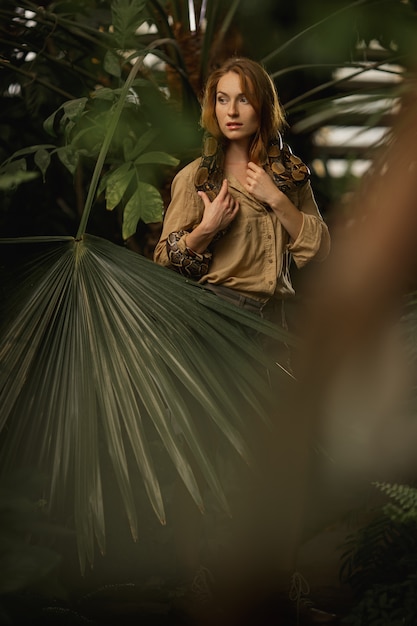 A beautiful girl with natural make-up and red hair stands in the jungle among exotic plants with snake.