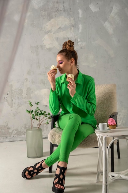 Beautiful girl with pink eyeshadow in studio on gray background Lady in green drinks coffee and eats muffins