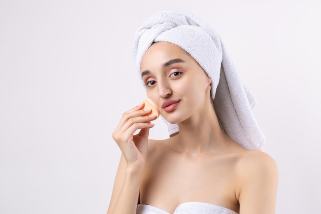 Beautiful girl with thick eyebrows and perfect skin at white background towel on head beauty photo Holds a cosmetic sponge