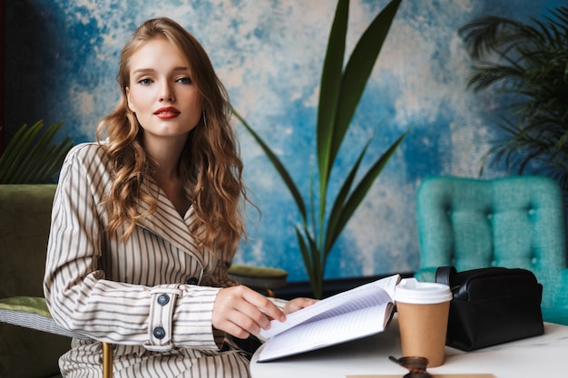 Beautiful girl with wavy hair in striped trench coat sitting with book in hands and coffee to go on table dreamily 