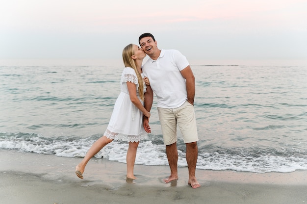 Beautiful girl with white hair kisses her husband on the cheek on the beach near the ocean at sunset.