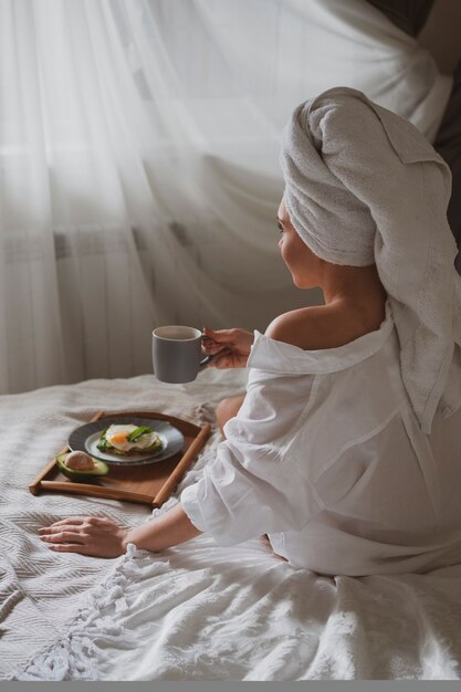 Beautiful girl with a white towel on her head sits in bed in the morning and drinks coffee