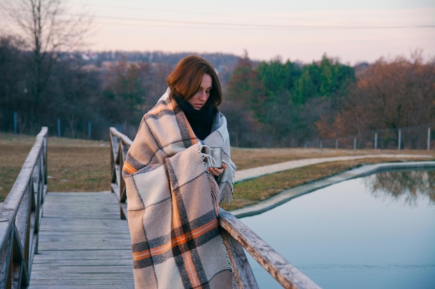 A beautiful girl wrapped in a plaid walks along the bridge over the pond with a metal mug of tea on a cold autumn day