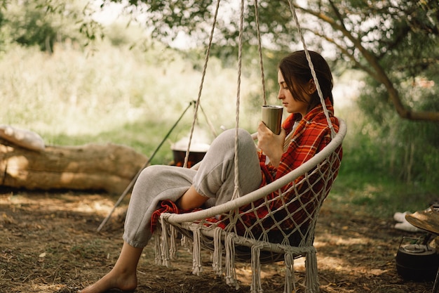 Beautiful girl wrapped in red plaid drinking tea in a cozy hanging chair outdoors adventure travel