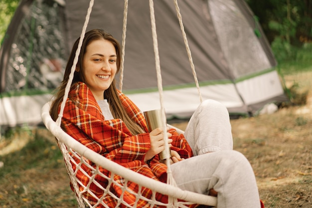 Beautiful girl wrapped in red plaid drinking tea in a cozy hanging chair outdoors adventure travel