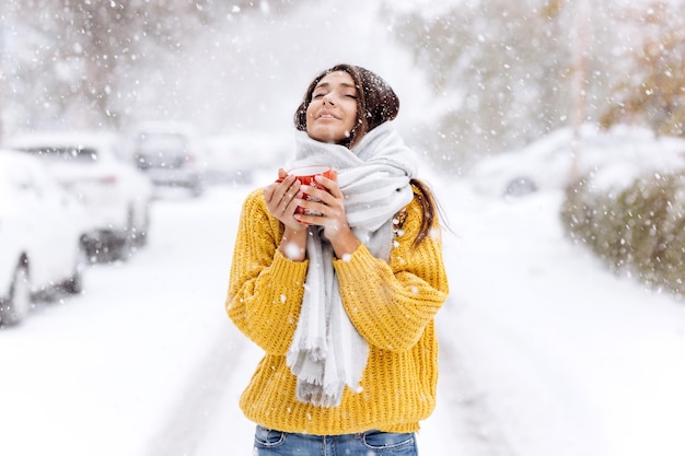 Beautiful girl in a yellow sweater and a white scarf standing with a red mug on a snowy street