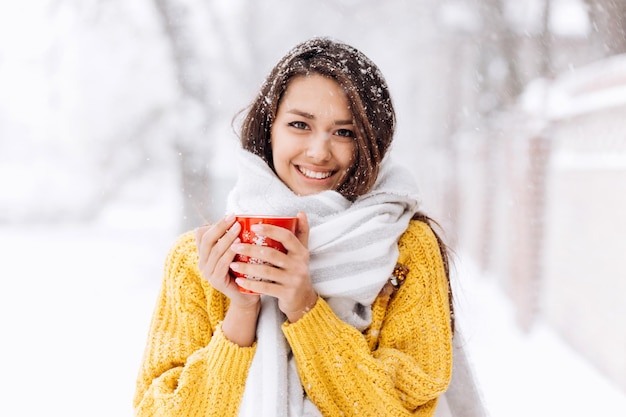 Beautiful girl in a yellow sweater and a white scarf standing with a red mug on a snowy street