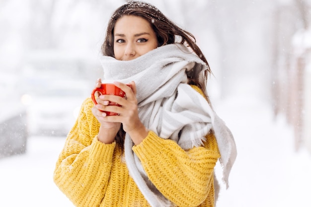 Beautiful girl in a yellow sweater and a white scarf standing with a red mug on a snowy street