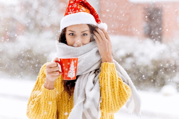 Beautiful girl in a yellow sweater and a white scarf standing with a red mug on a snowy street