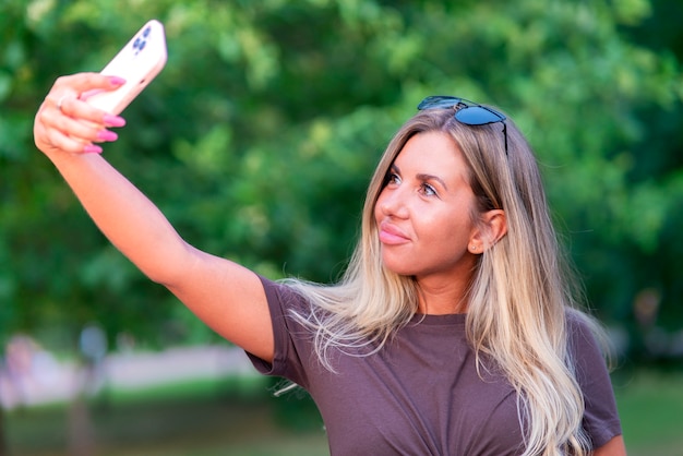 Beautiful girl, young happy sexy woman looking at camera, taking a photo, picture of herself, selfie on cell phone, modern smartphone, smiling in summer park. Mobile photographing