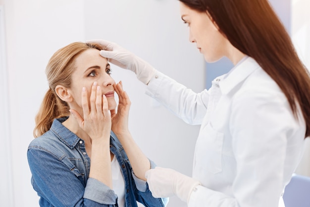 Beautiful good looking nice woman holding her cheeks and looking at the doctor while wanting to do a plastic surgery