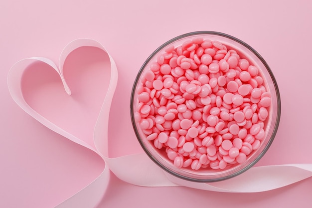 Beautiful granules of pink wax for depilation in a glass bowl and a ribbon in the shape of a heart