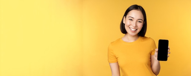 Beautiful happy asian girl showing mobile phone screen application on smartphone gadget standing over yellow background