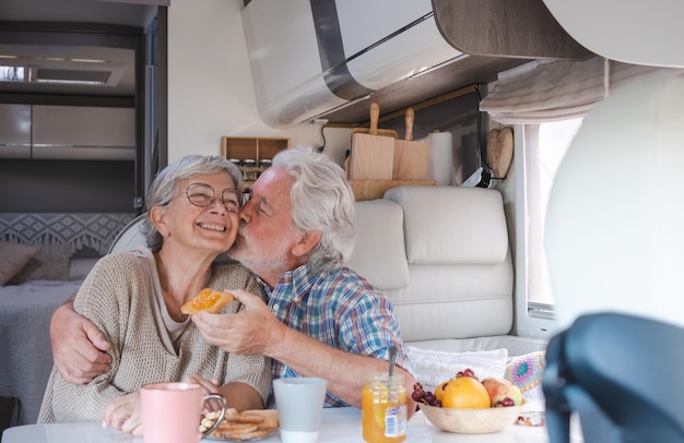 Beautiful happy caucasian senior couple in travel vacation sitting inside a camper van dinette enjoying breakfast together husband kisses his wife