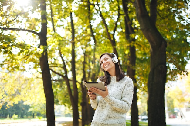 The beautiful happy cheerful brown-haired woman in white sweater with a tablet listening music in the white headphones in fall park on a warm day. Autumn in the city.